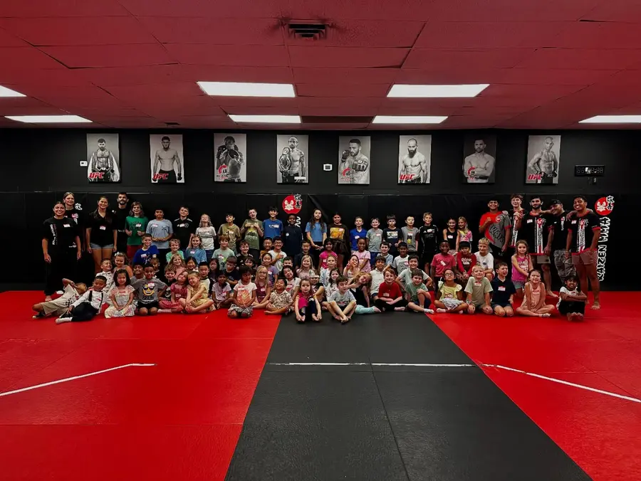 A wide training room with children sitting along the edge of the mats